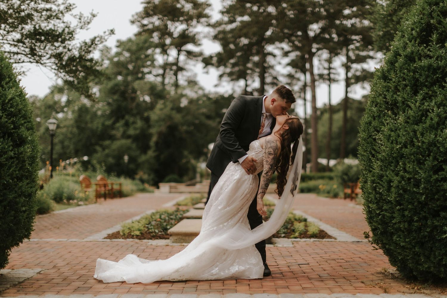 Wedding couple poses on brick pathways surrounded by manicured gardens and trees.