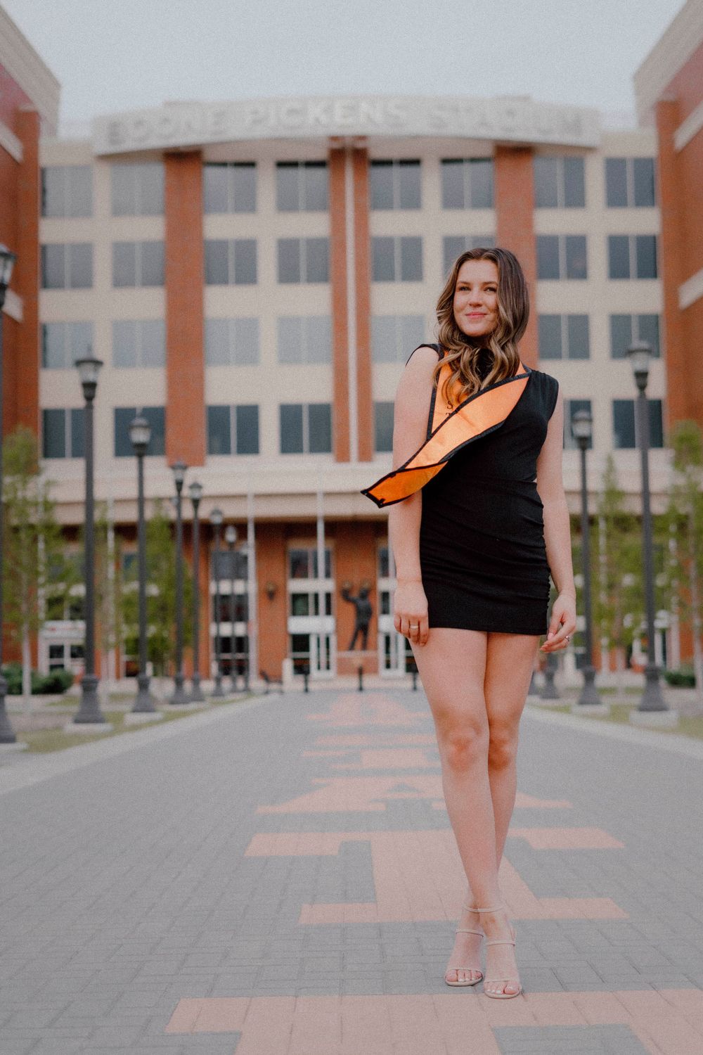 Graduate in black dress and orange stole poses on campus walkway with building in background.