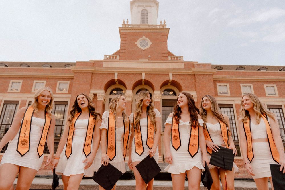 Graduates in white dresses and orange stoles pose on steps of campus building with bell tower.