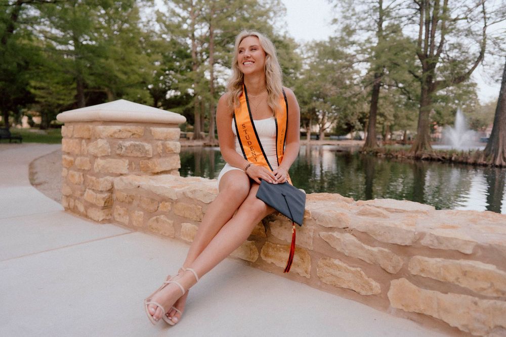 Graduate with orange stole sits on stone wall near pond during sunset.