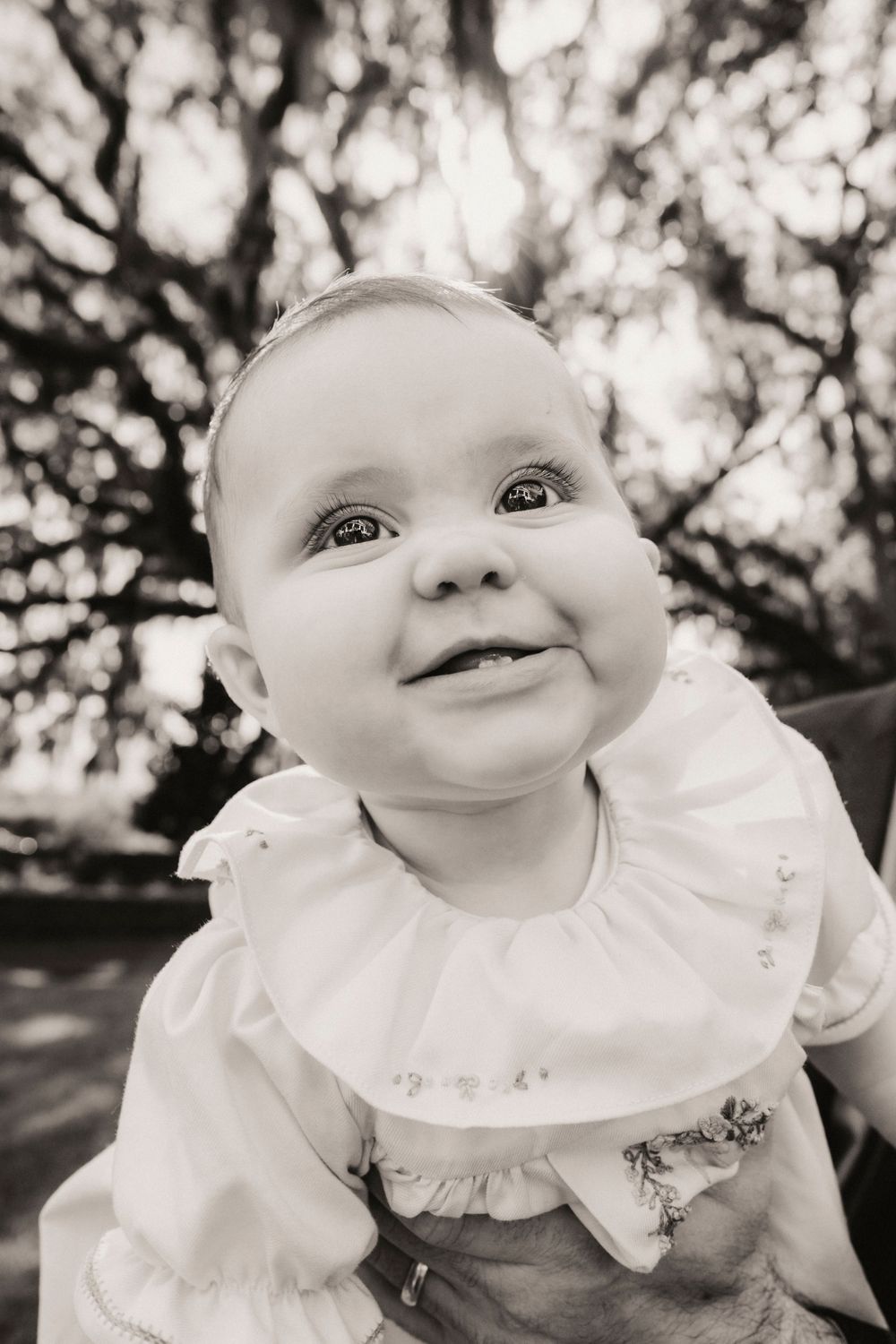 A baby wearing a white ruffled dress smiles at the camera in an outdoor garden setting.