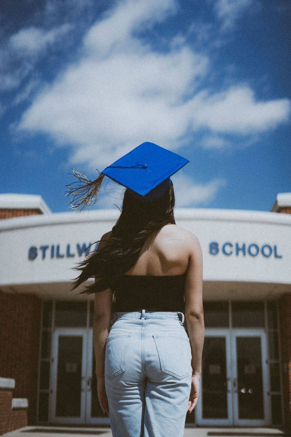 A graduate in light denim faces away from camera in front of Stillwater High School entrance.