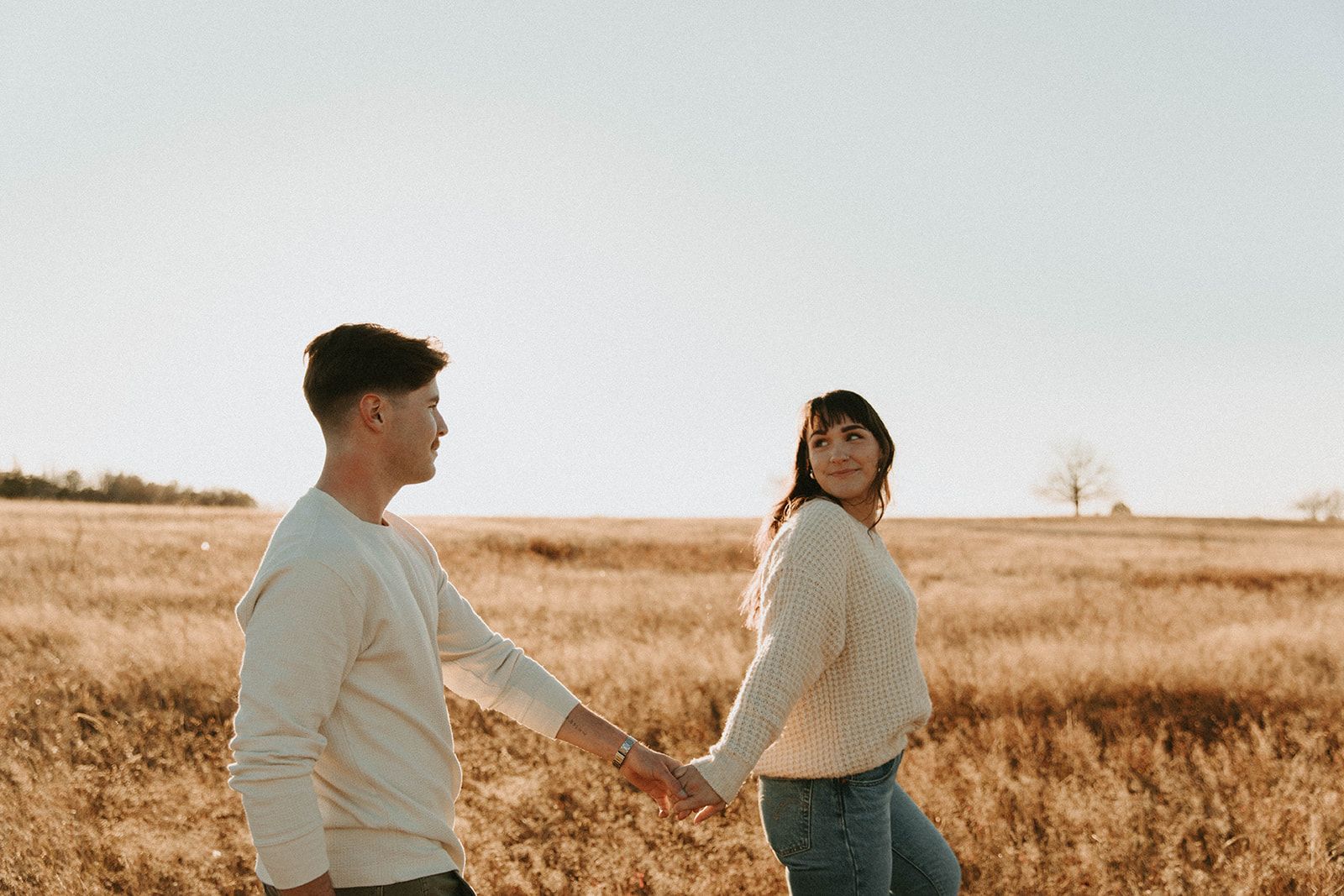 Couple in matching cream sweaters holding hands while walking through a golden autumn field.
