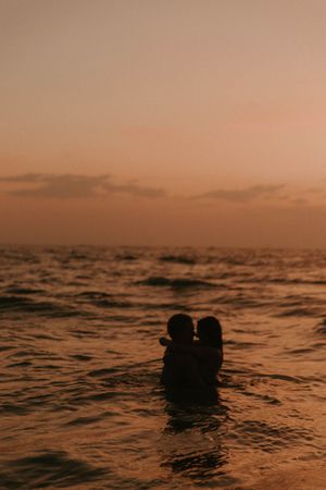 Two silhouettes embrace in ocean waters during a golden sunset.