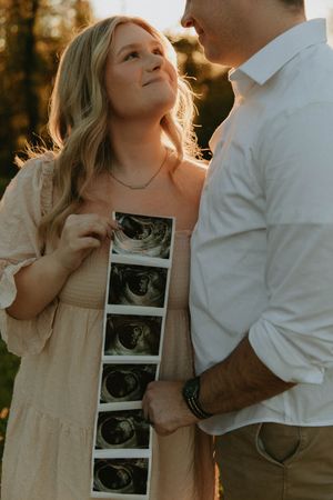 A couple shares a loving moment while holding ultrasound photos outdoors.