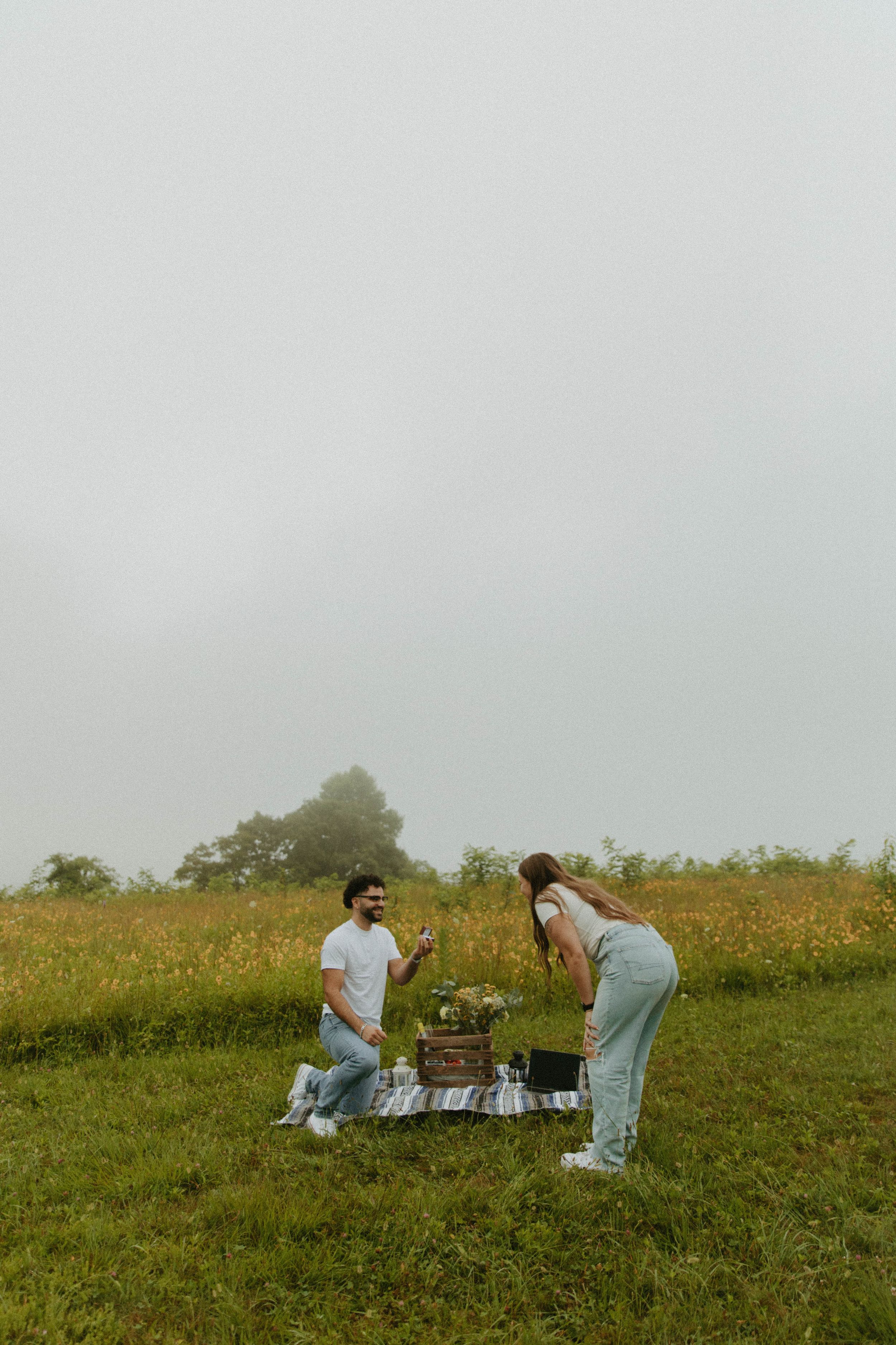 A couple setting up a romantic picnic on a grassy hilltop during a misty day.