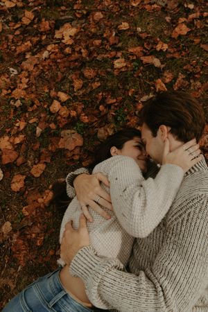 A couple in cozy sweaters shares an intimate moment while lying on fallen autumn leaves.