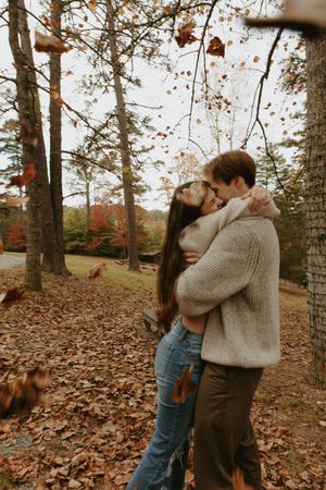 A romantic couple embraces among fall foliage and tall trees on an autumn day.
