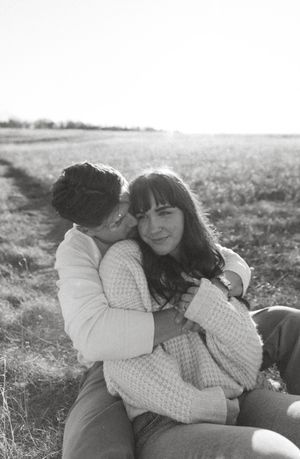 A couple in matching white sweaters embraces on a grassy field in black and white photography.