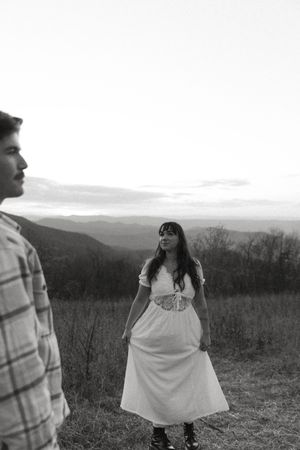A couple stands in a mountainous field at sunset with the woman wearing a flowing white dress.