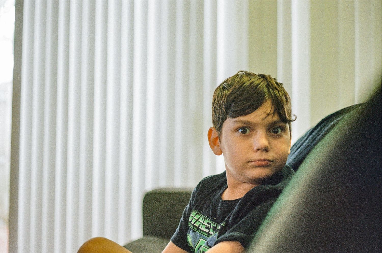 Youth lounging on couch near vertical blinds in natural light.