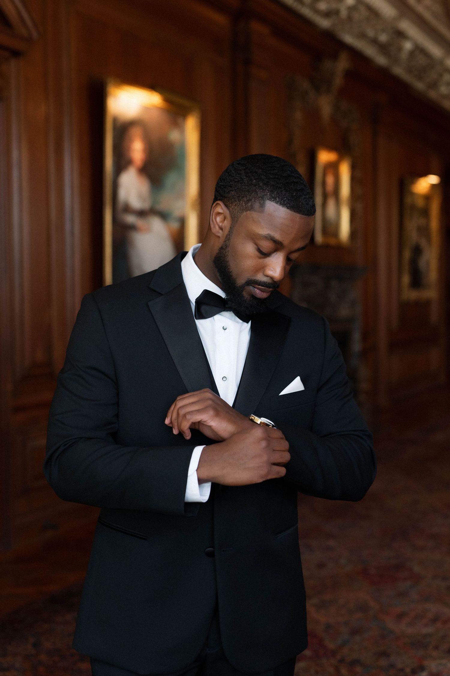 Groom in classic black tuxedo adjusting cufflinks in wood-paneled room.