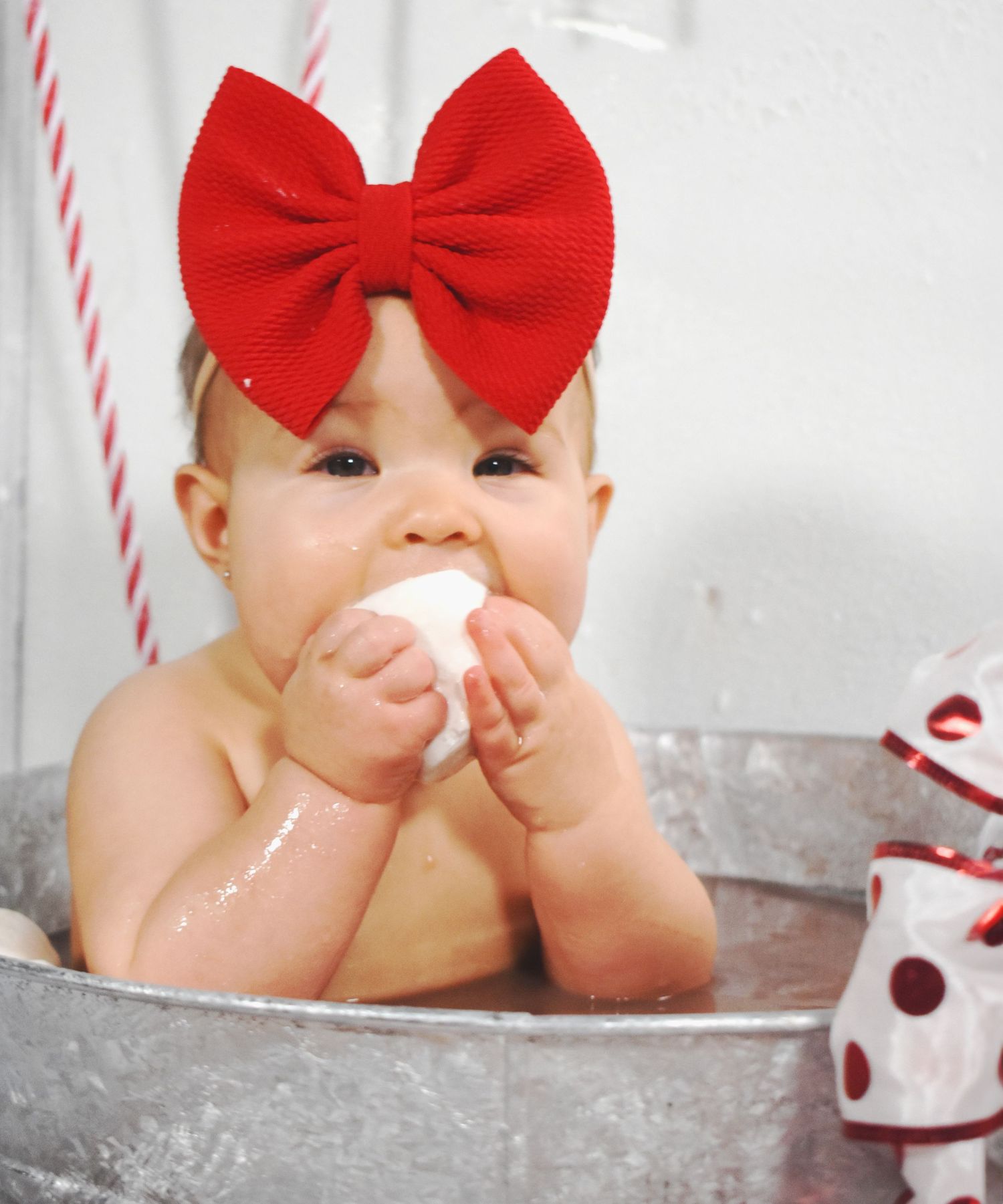 Baby in red bow playing with marshmallows in metal tub during holiday photoshoot.