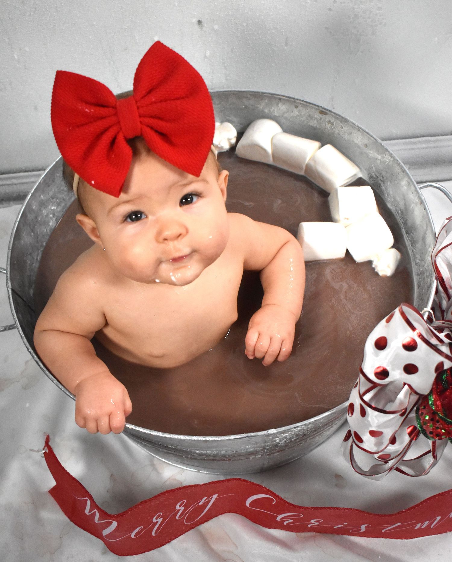 Baby wearing red bow sits in galvanized tub surrounded by Christmas decorations.