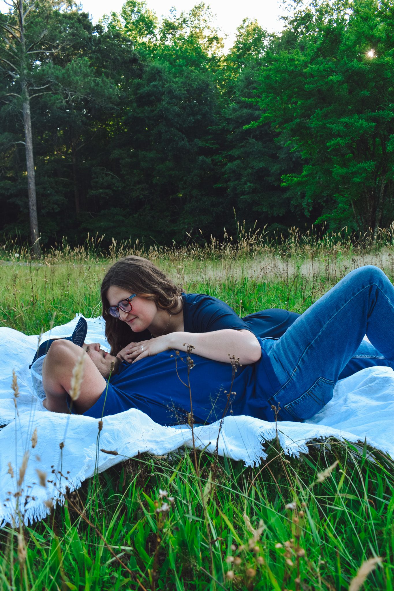 People relaxing on a white blanket in a sunny meadow surrounded by trees.