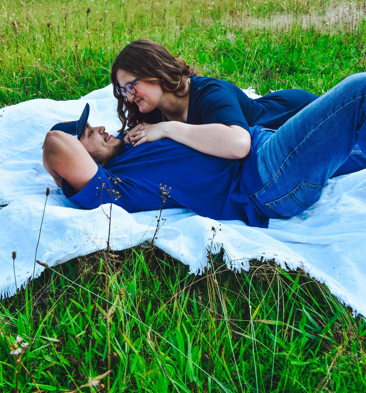 A romantic couple lying on a white blanket in green grass sharing an intimate moment together.