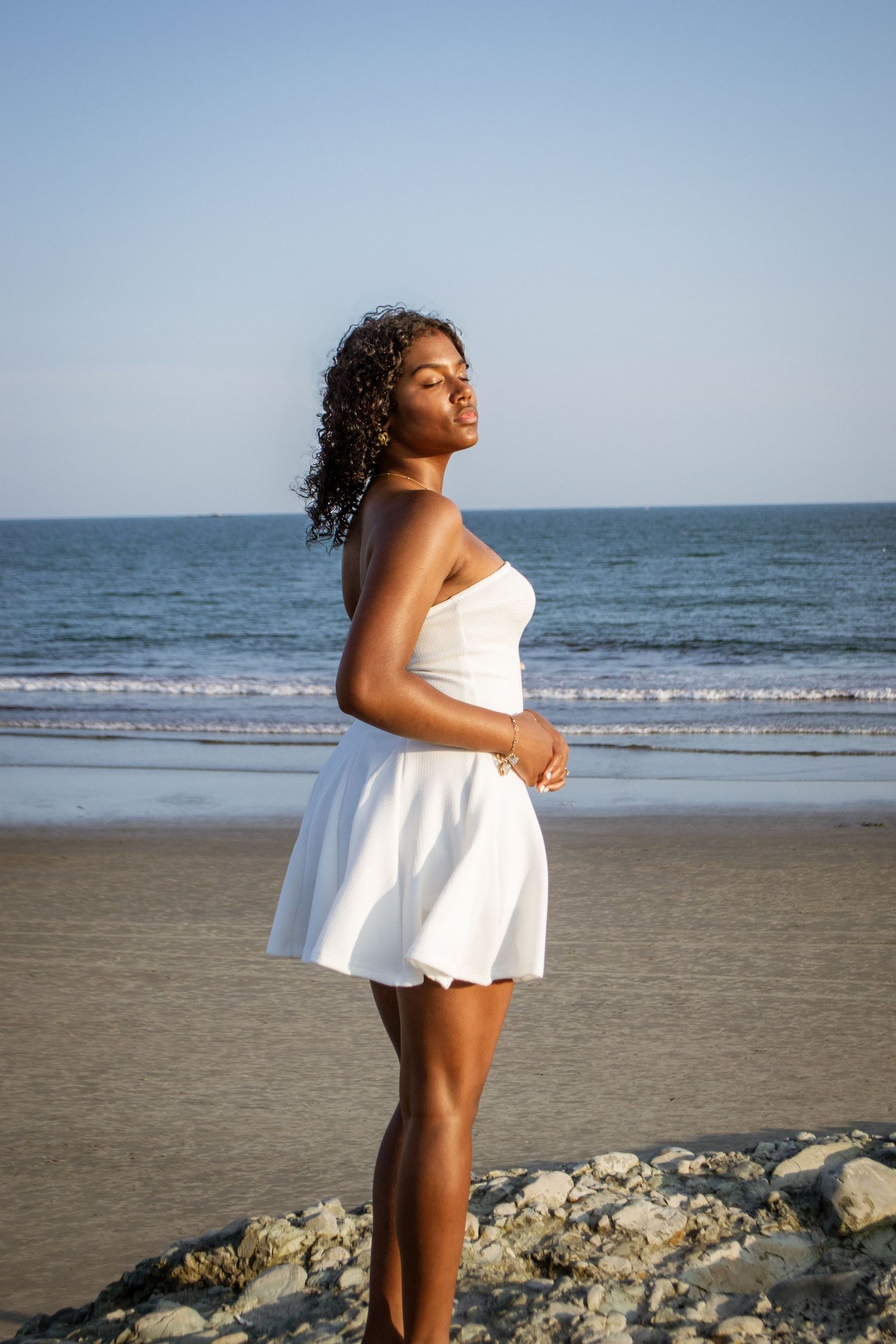 Person in white dress stands confidently against ocean backdrop.