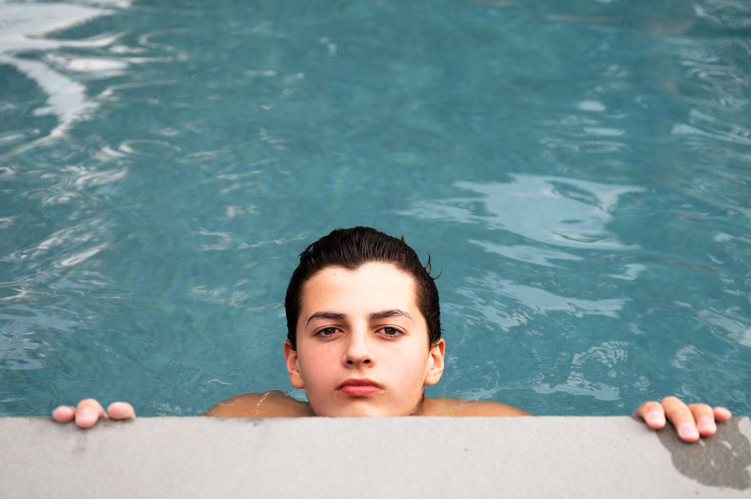 A person holding onto the edge of a pool with turquoise blue water in the background.