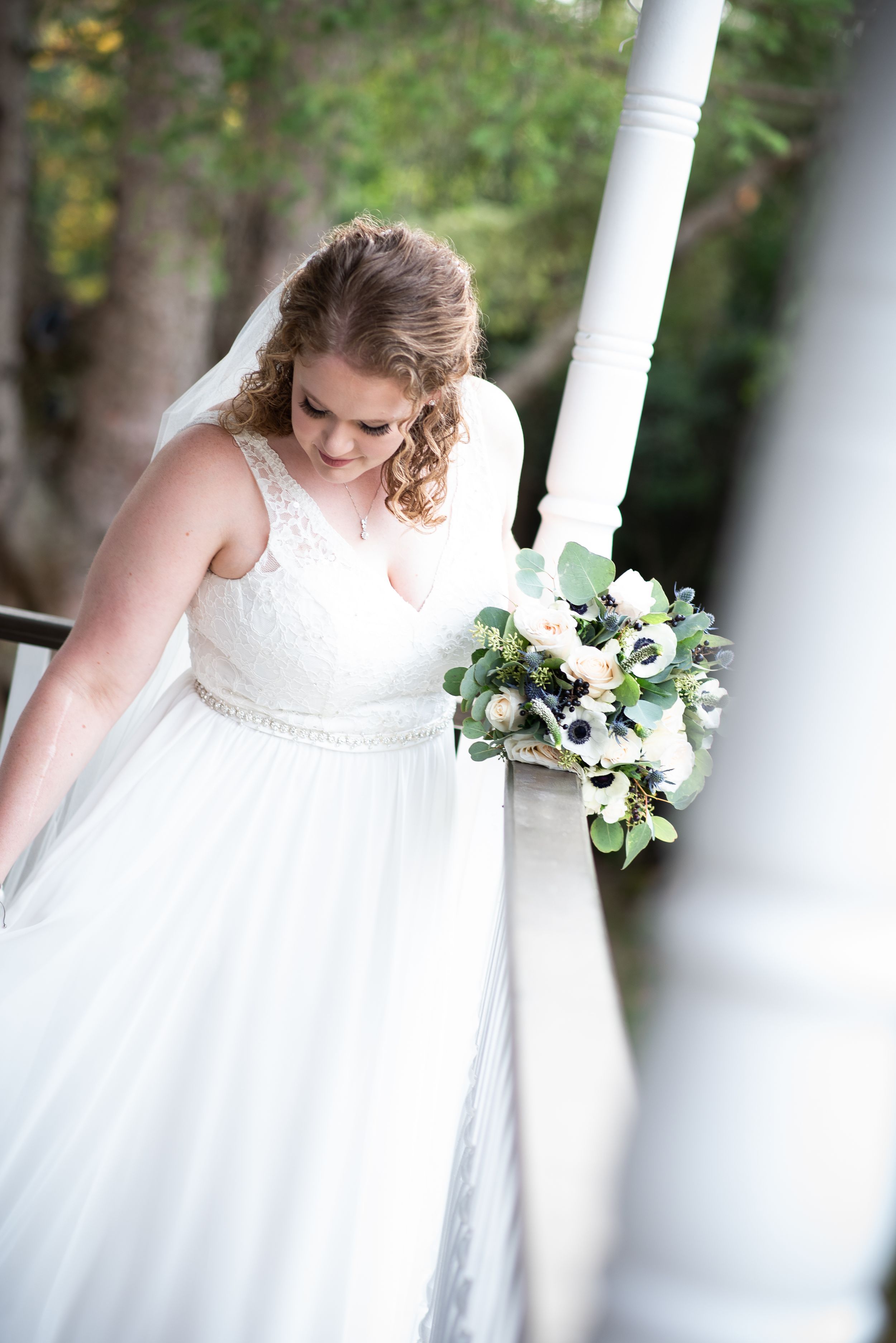 A bride in a flowing white gown with beaded details holds a soft pastel bouquet while standing on a white columned porch.