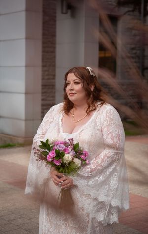 A bride in a white lace wedding dress poses with a pink and white floral bouquet against a brick building.