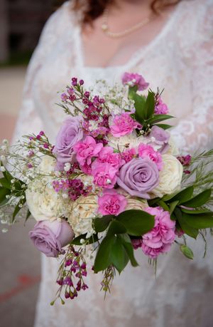 A beautiful wedding bouquet featuring pink carnations, purple roses, white hydrangeas, and delicate greenery.