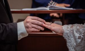Two hands exchanging a ring during a wedding ceremony at a wooden table.