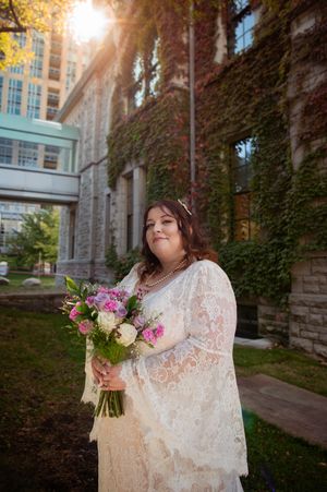 A bride in a vintage-style lace gown holds a colorful bouquet against an ivy-covered stone building at sunset.