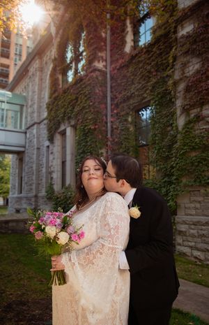 A romantic wedding couple embraces in front of an ivy-covered stone building during golden hour with a floral bouquet.
