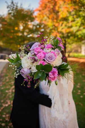 Beautiful wedding bouquet with purple roses, pink carnations and white flowers against autumn foliage background.