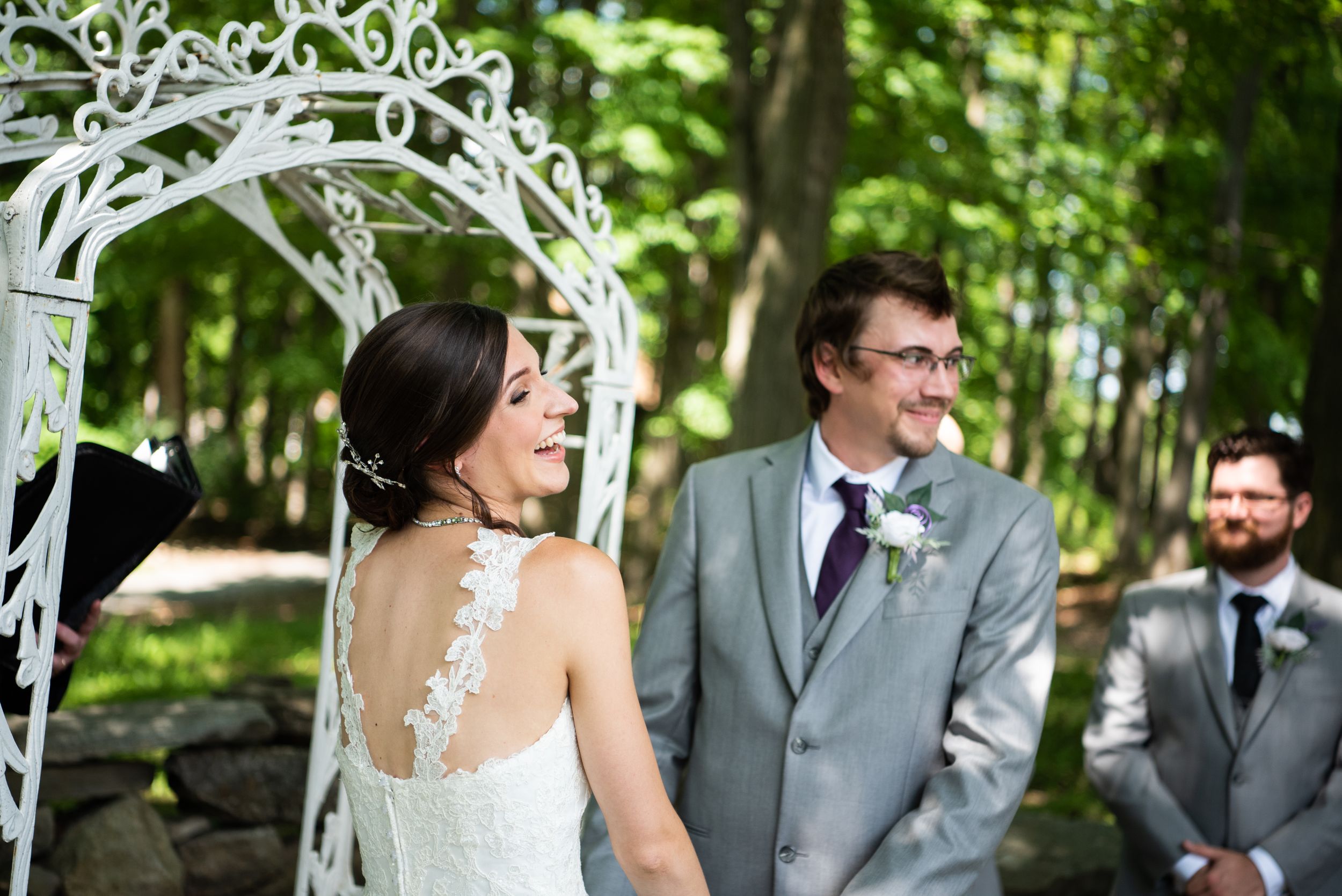 A wedding ceremony taking place under a white decorative arch in a lush green garden setting.