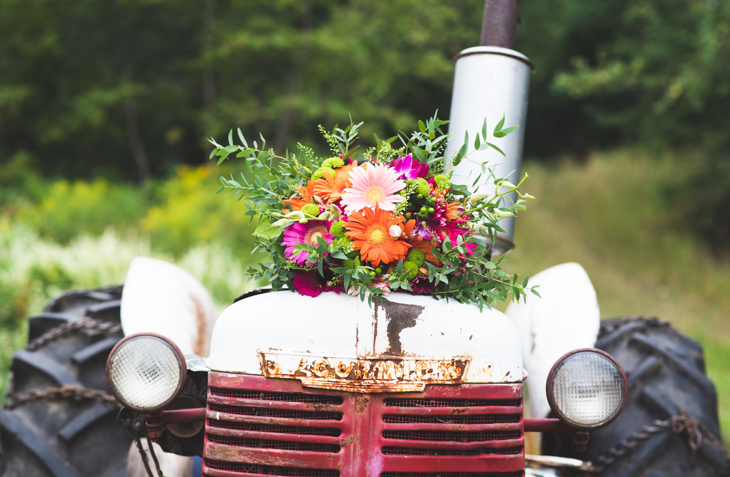 A rustic floral arrangement sits on the hood of a vintage tractor with large tires in an outdoor setting.