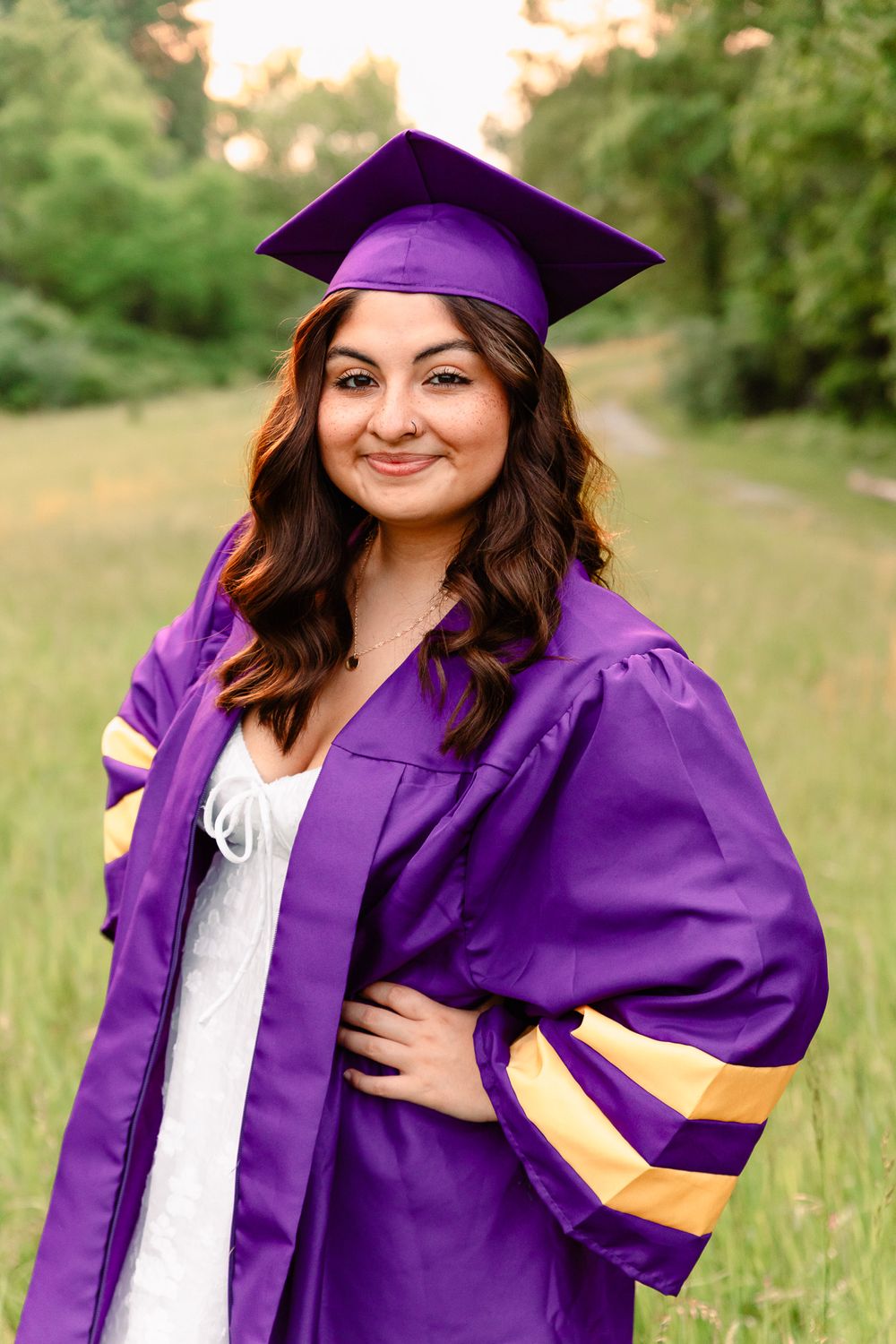 Graduate poses in purple graduation cap and gown in an outdoor field setting.