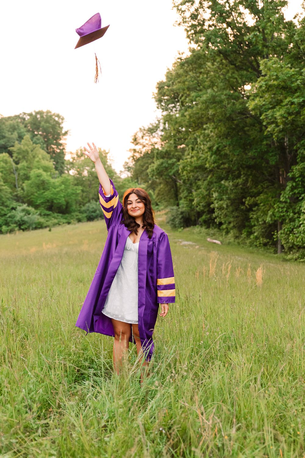 Graduate in purple regalia walking through tall grass during sunset photoshoot.