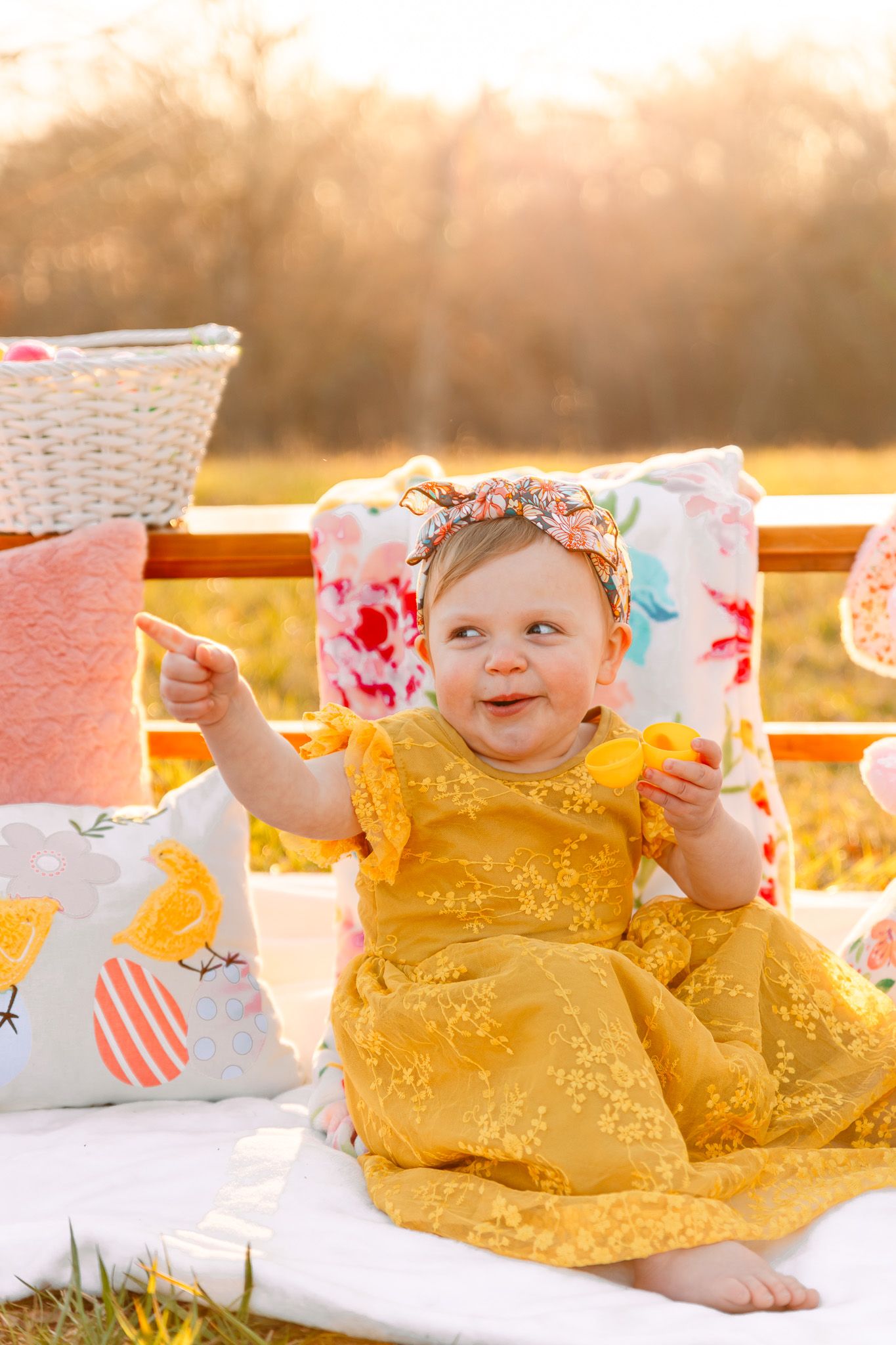 Baby wearing yellow dress sits among floral decorations during sunset photo session.