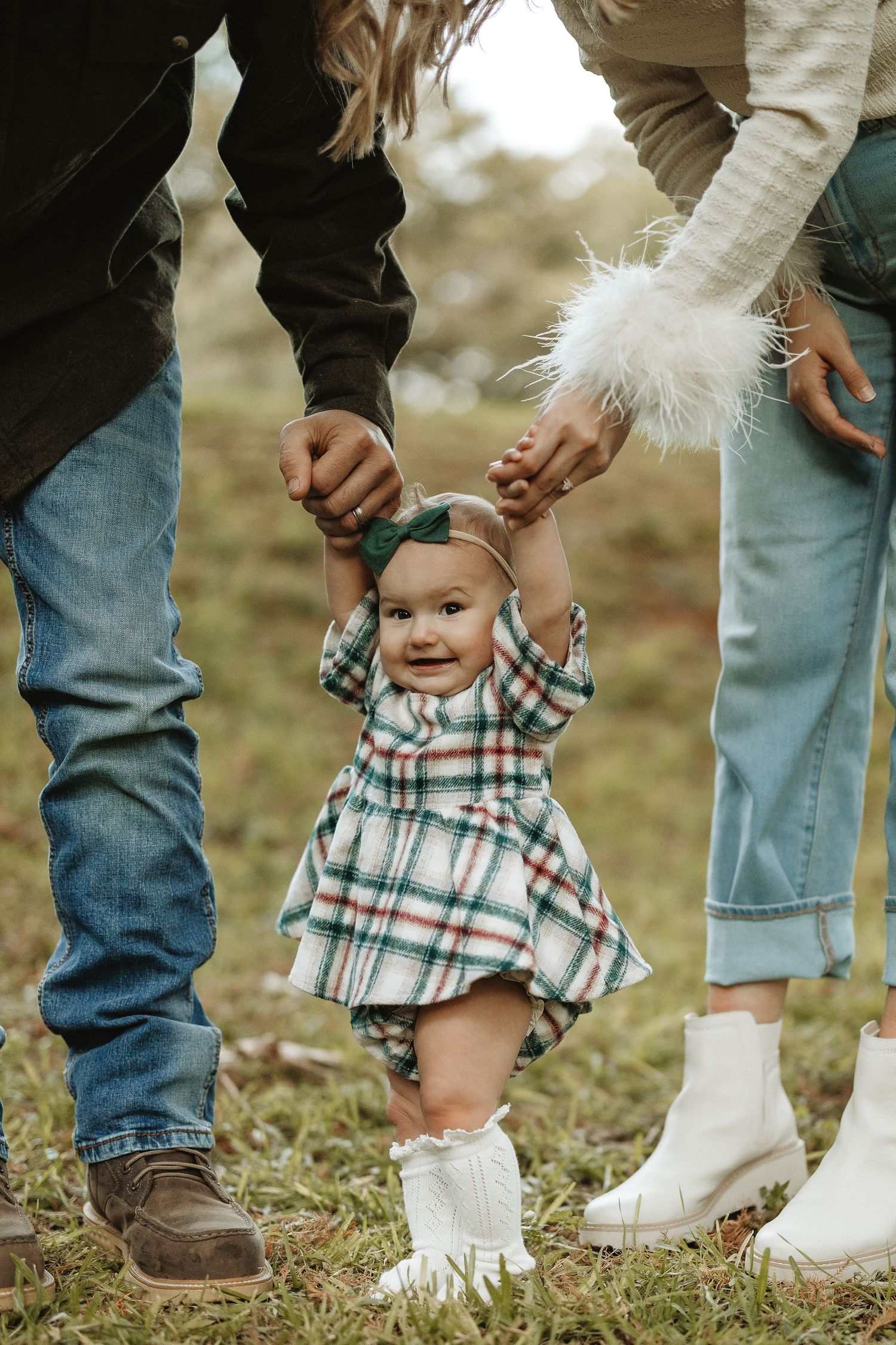 Small child in plaid dress and white boots standing between two people in jeans on grass.