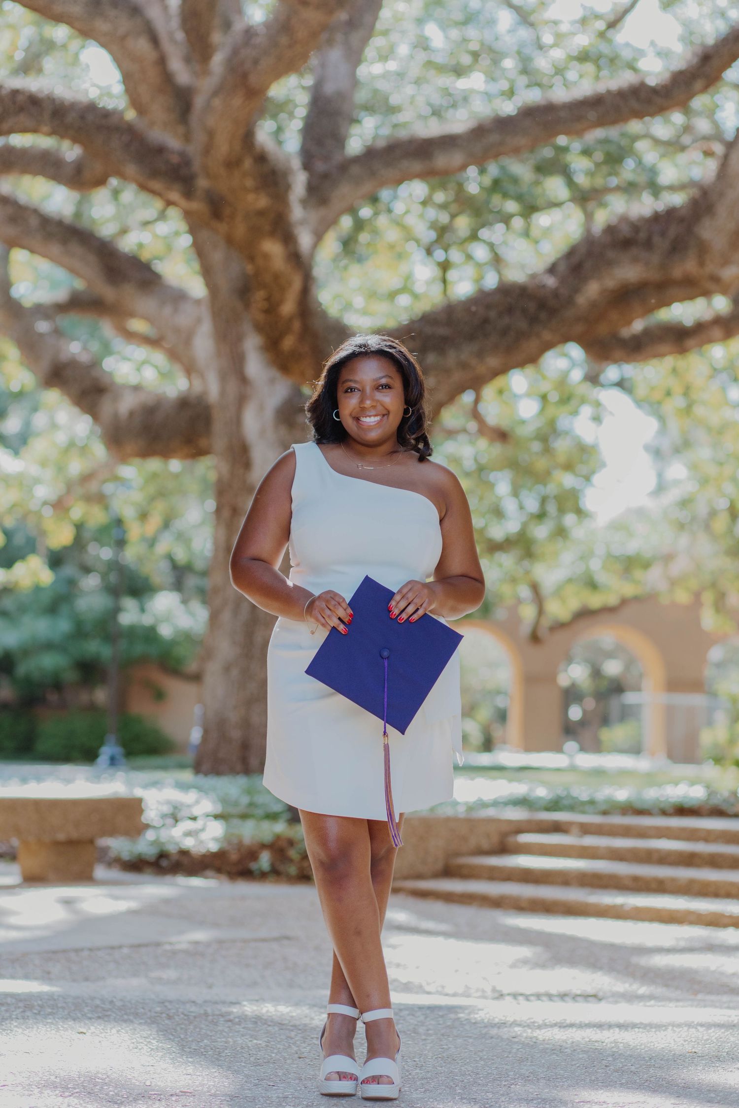 Graduate in a white one-shoulder dress poses confidently with diploma under a majestic oak tree.