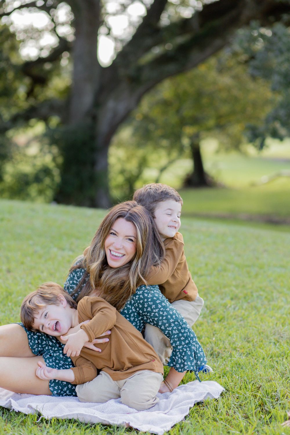 Family sitting together on a picnic blanket in a park with a large tree in the background on a sunny day.
