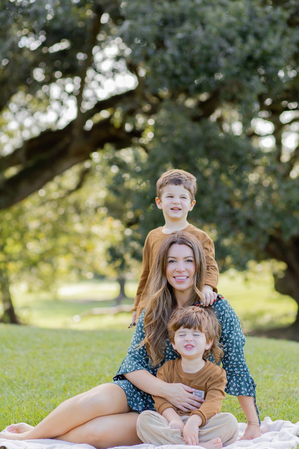 Family sits together on blanket outdoors enjoying sunny day under large tree in park setting.