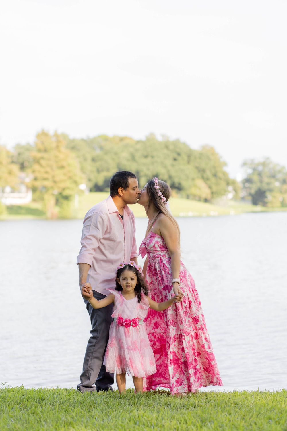 Family in coordinating pink outfits poses by a lakeside on a sunny afternoon.