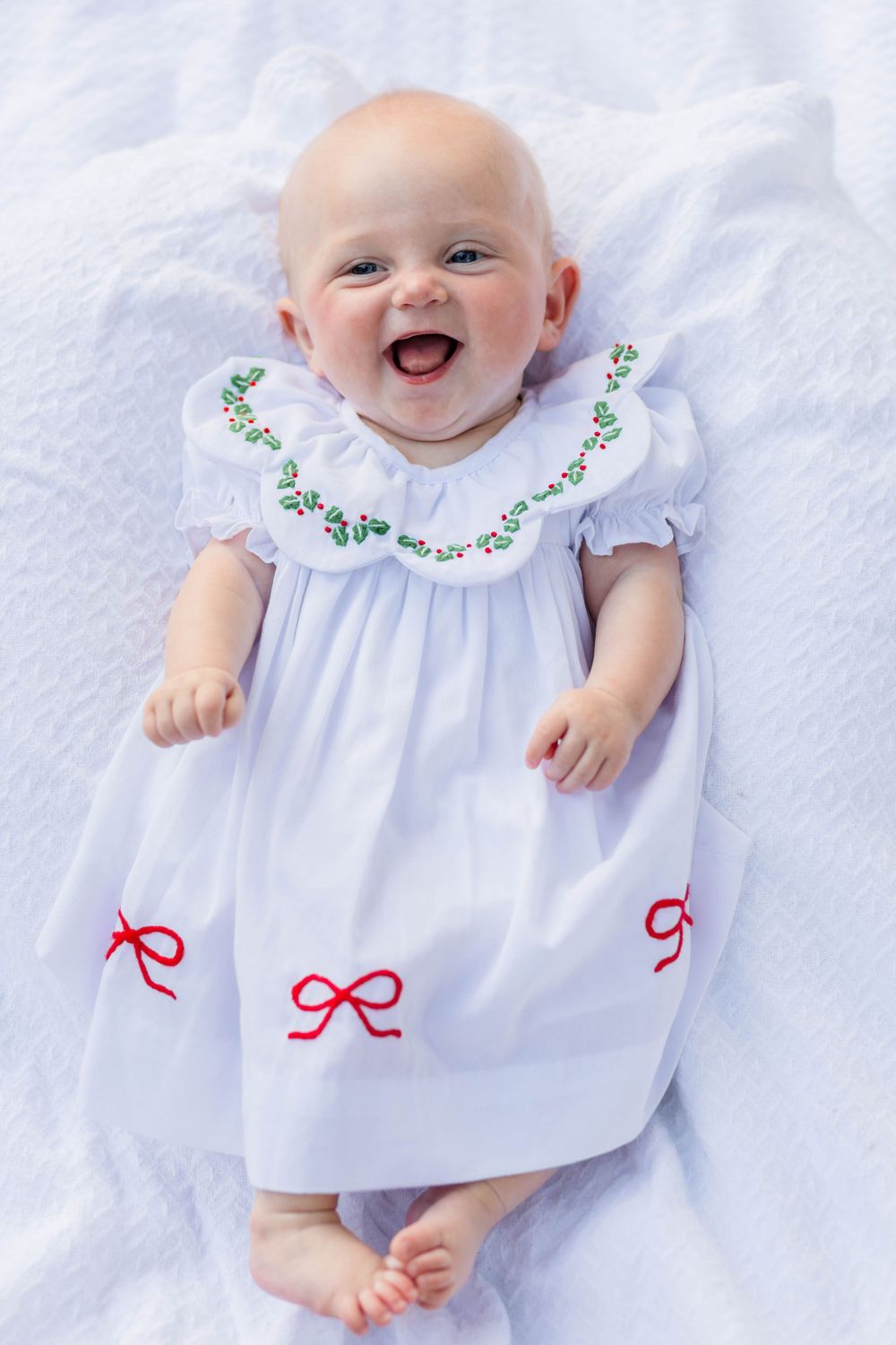 Baby wearing white dress with green floral embroidery and red bow details lies on white fabric while smiling joyfully.