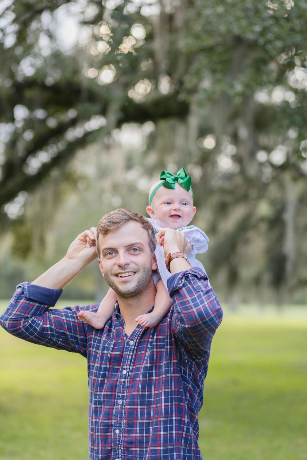 A young child wearing a green bow sits on adult shoulders outdoors with trees in the background.