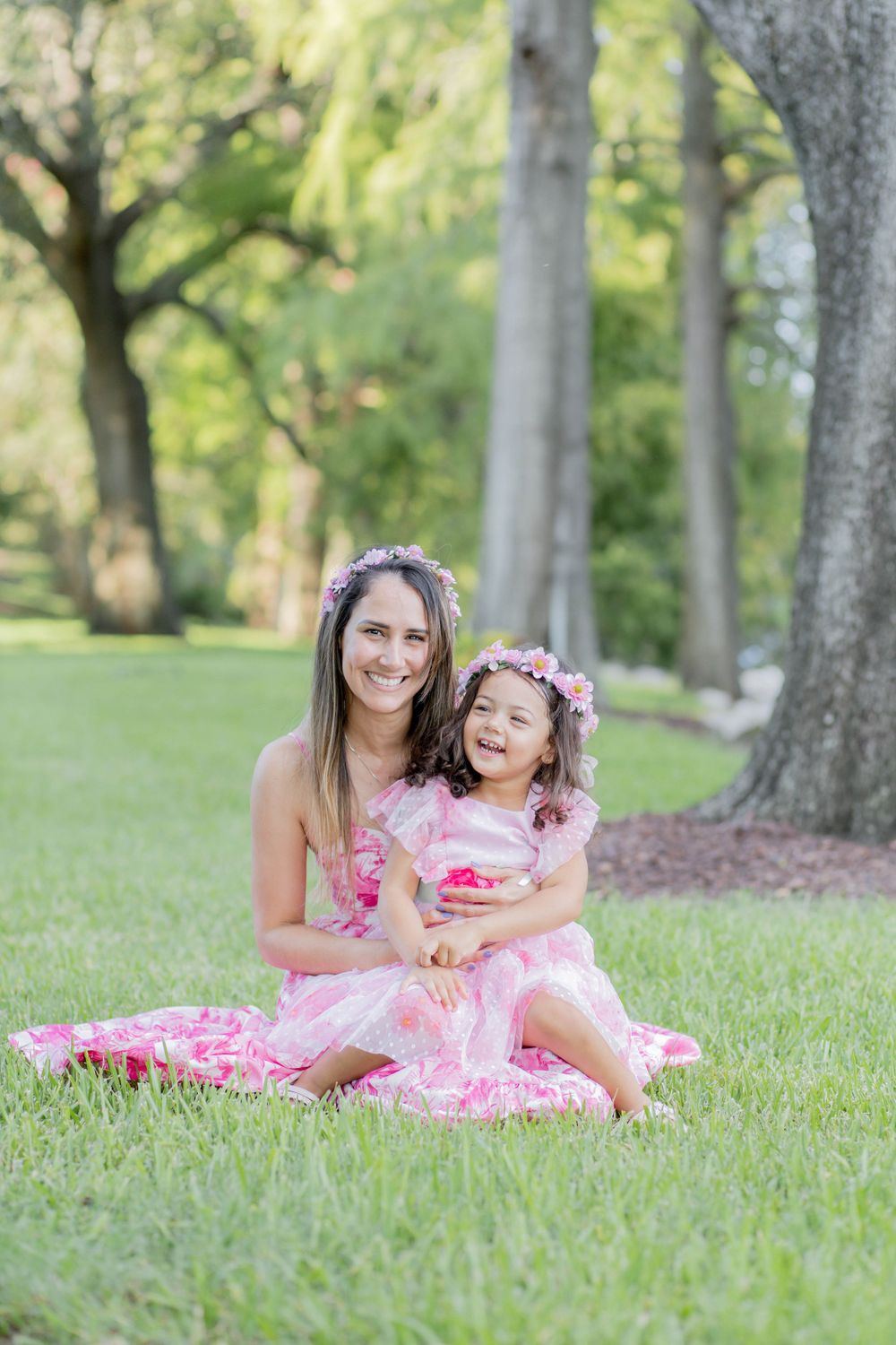 Two people in pink outfits share a tender moment while sitting on grass in a park setting.