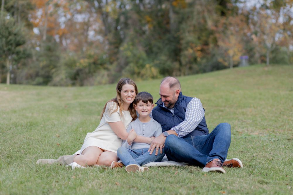 A family sits together on a blanket on grass during autumn with trees in the background.