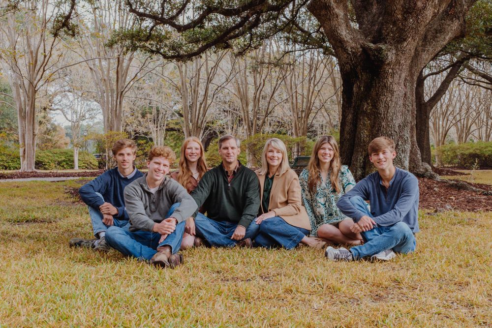 Large family group sits casually on grass for outdoor portrait under sprawling oak trees.