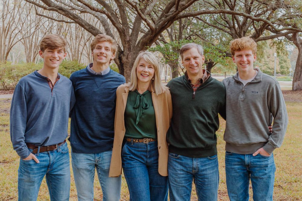 Group of five people in casual attire pose together for outdoor portrait against bare trees.