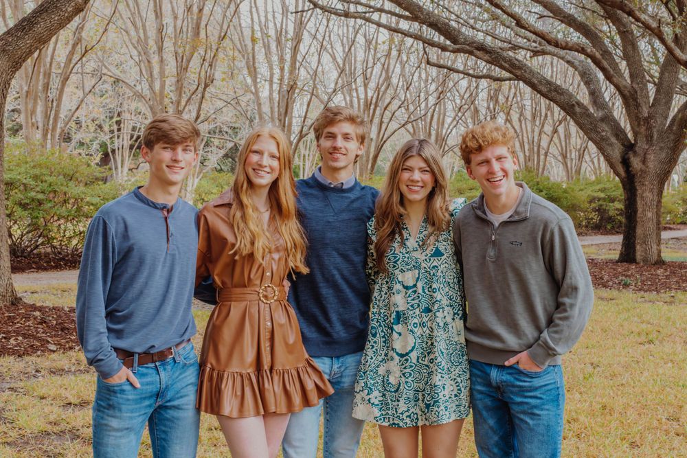 Five people in coordinated casual outfits pose for outdoor family portrait among trees.