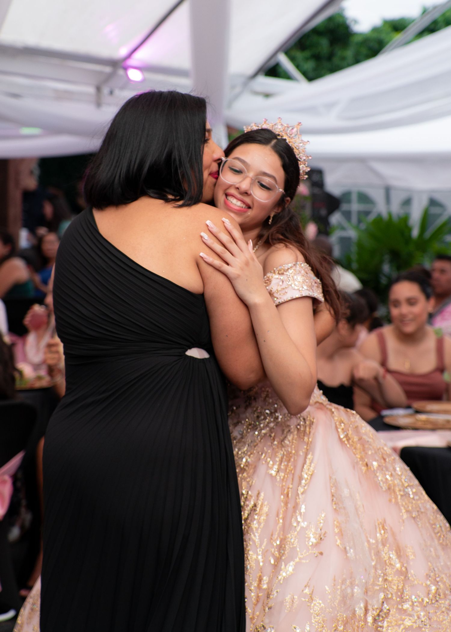 Woman in black dress hugging a smiling girl in a pink sequin quinceañera gown at a celebration.
