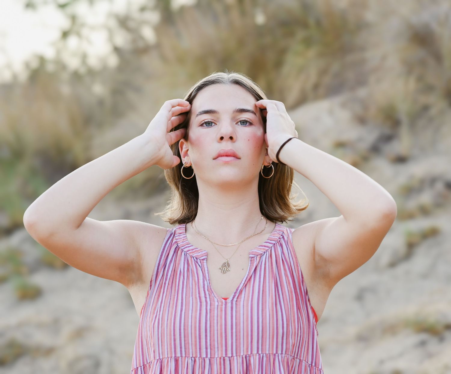 Woman in striped pink dress poses with hands in hair against natural outdoor setting.