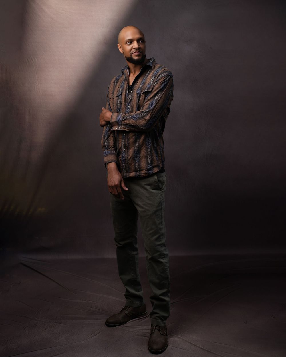 Studio portrait of someone in a striped casual shirt and dark pants standing against a dramatic shadowy background.