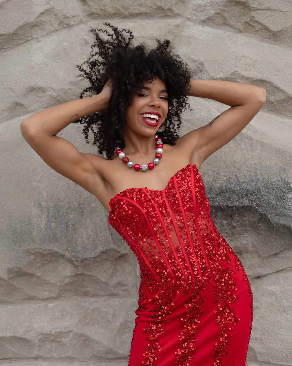 Joyful model in a sparkling red strapless gown poses with hands in curly hair against a textured stone wall.